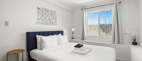 A modern hotel room with a large bed, a bedside table, a wall art piece, a window with curtains, and a view of the outdoors.