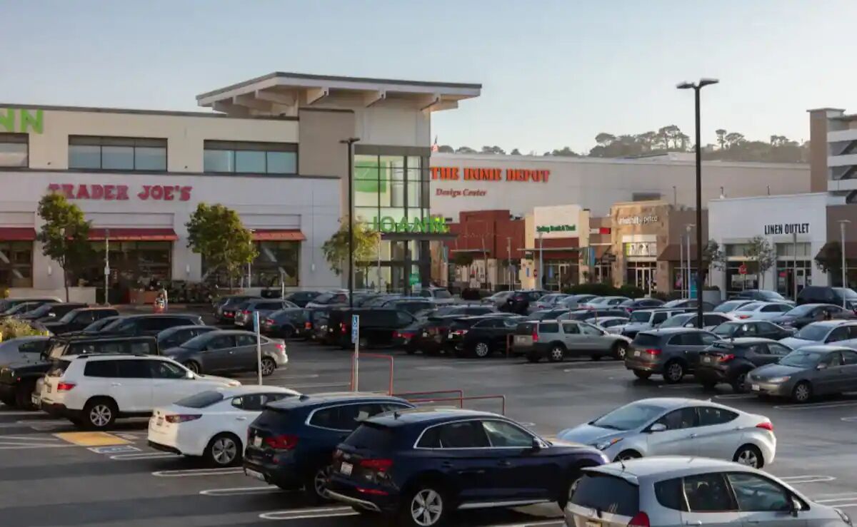 Un parking rempli de voitures, avec un centre commercial en arrière-plan.