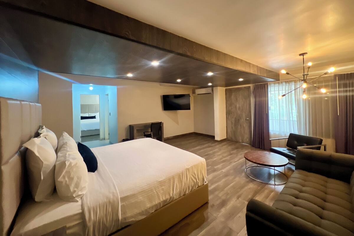A modern hotel room with a large bed, a sofa, a TV, and a dining area.