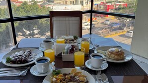 A breakfast table with plates of food, cups of coffee, and glasses of orange juice, viewed through a large window overlooking a cityscape with buildings and construction. - Hotel Sunio City (Tijuana)