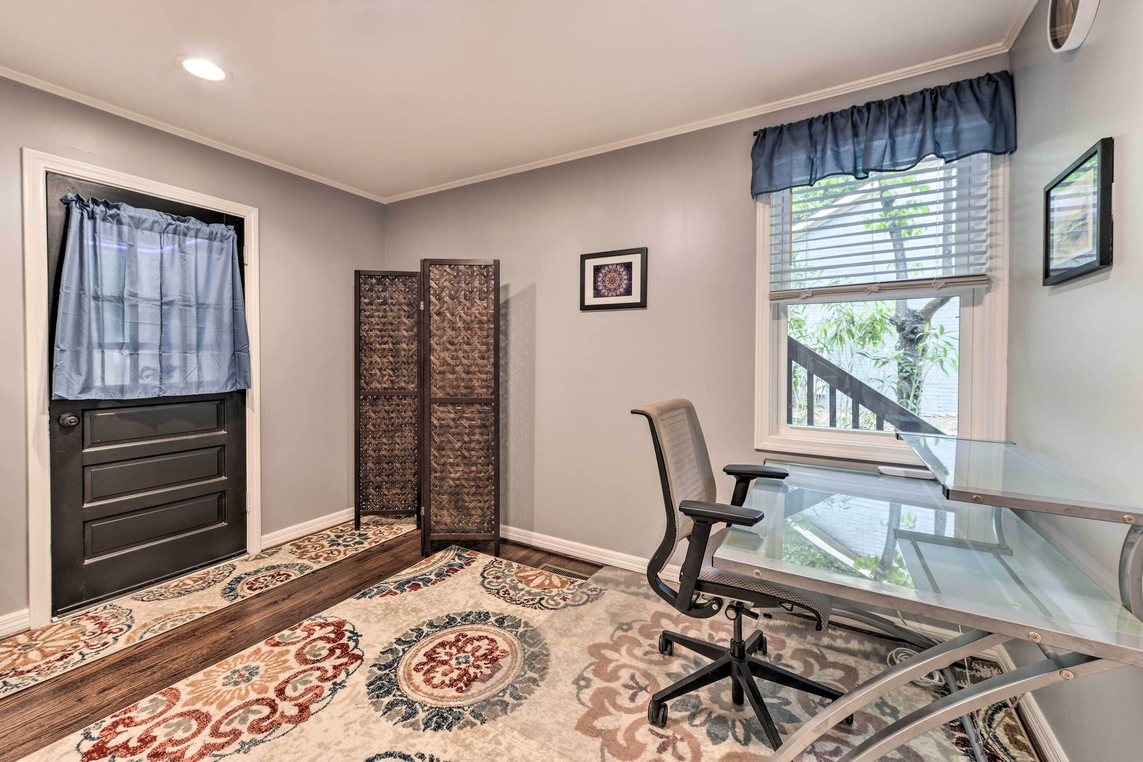 Une pièce avec une porte sombre, un tapis à motifs, un bureau en verre avec une chaise, une fenêtre avec des stores et un tableau encadré au mur.