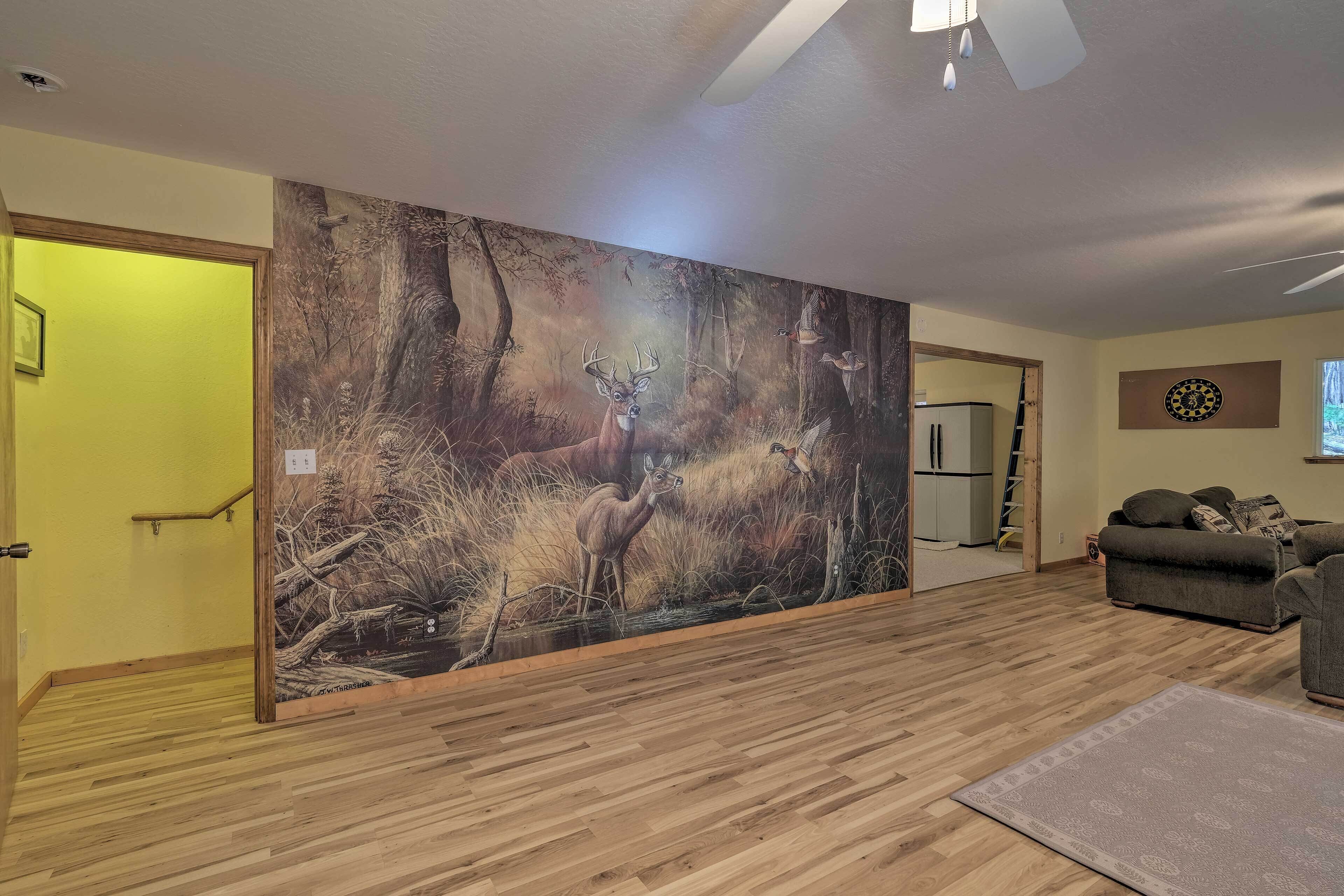 A living room with a large deer mural on the wall, wooden flooring, a gray sofa, and a dartboard.