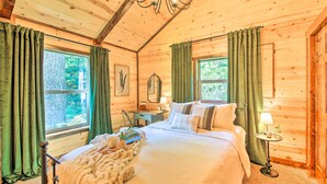 A wooden cabin bedroom with a bed, a nightstand, a lamp, and a window with curtains. - Rustic-chic Woodland Hideaway w/ Hot Tub! (Sandpoint)