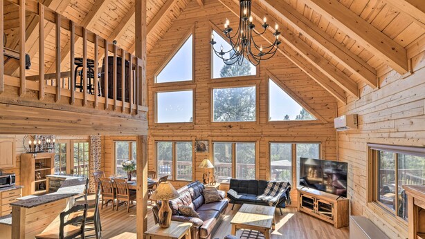 A spacious log cabin interior with a high ceiling, large windows, a dining area, a living room with a TV, and a fireplace. - Lazy Beaver Lodge Pagosa Cabin w/ Deck, Fire Pit (Pagosa Springs)