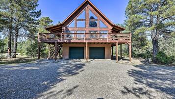 Une cabane en rondins à deux étages, dotée d’un balcon, d’un garage et d’une allée en gravier, le tout entouré d’arbres.
