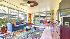 A spacious living room with a blue sofa, a glass coffee table, and a colorful rug. There is a large window offering a view of palm trees and a garden. - Island Oasis Near Kaanapali Beach & Black Rock (Kaanapali)