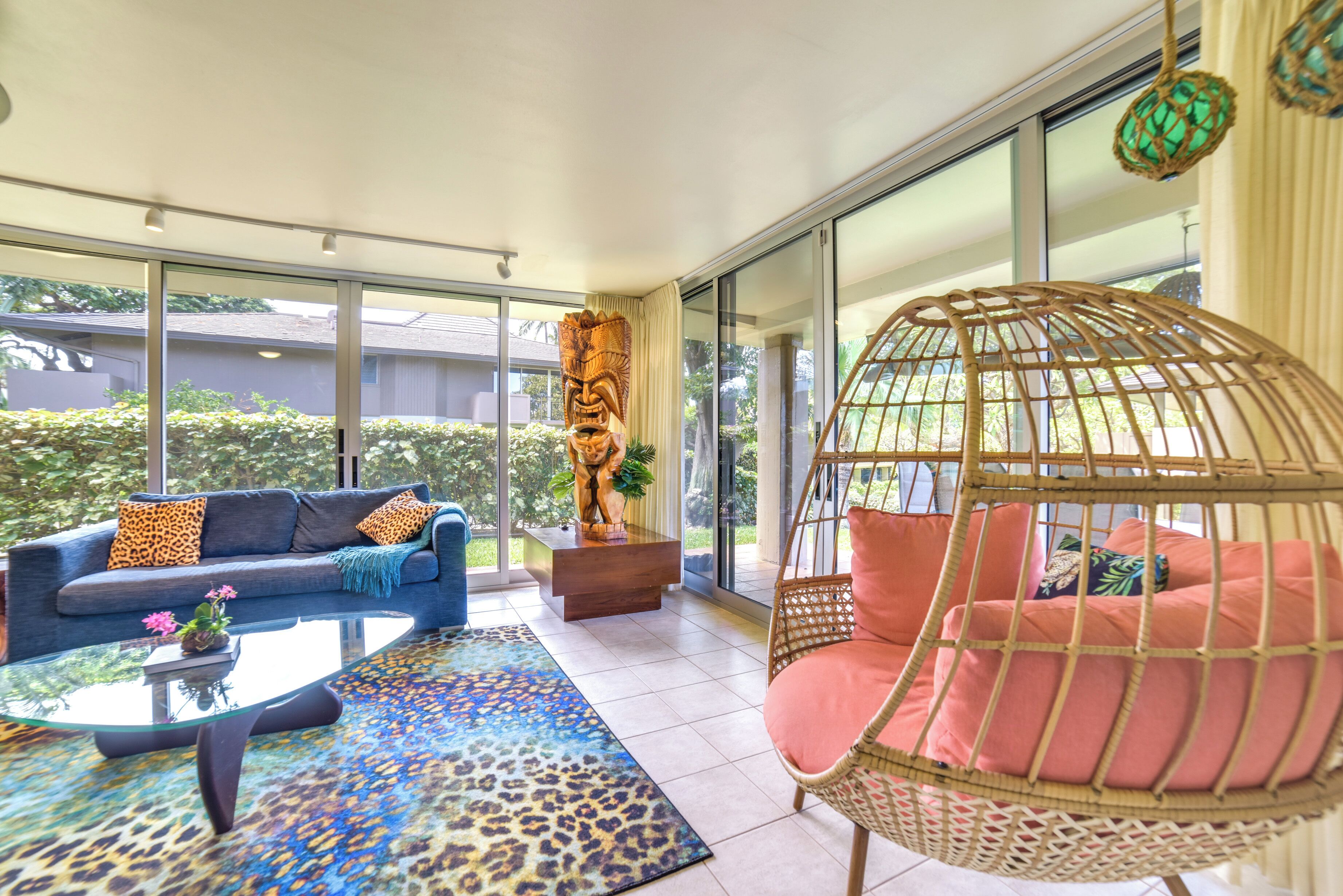 Ein modernes Wohnzimmer mit einem Glastisch, einem blauen Sofa mit Leopardenmuster-Kissen, einem Korbstuhl mit pinken Kissen und einer Statue auf einer Holzplattform.