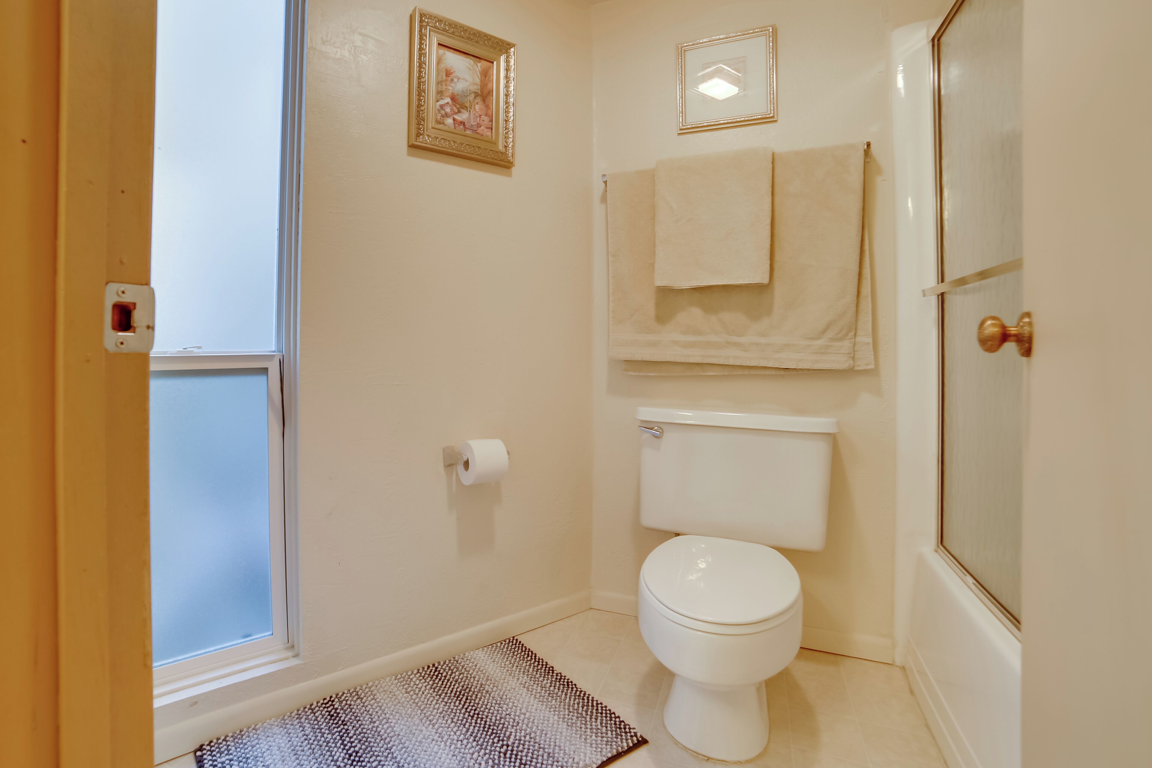 A bathroom with a toilet, a towel rack, a framed picture, and a window.