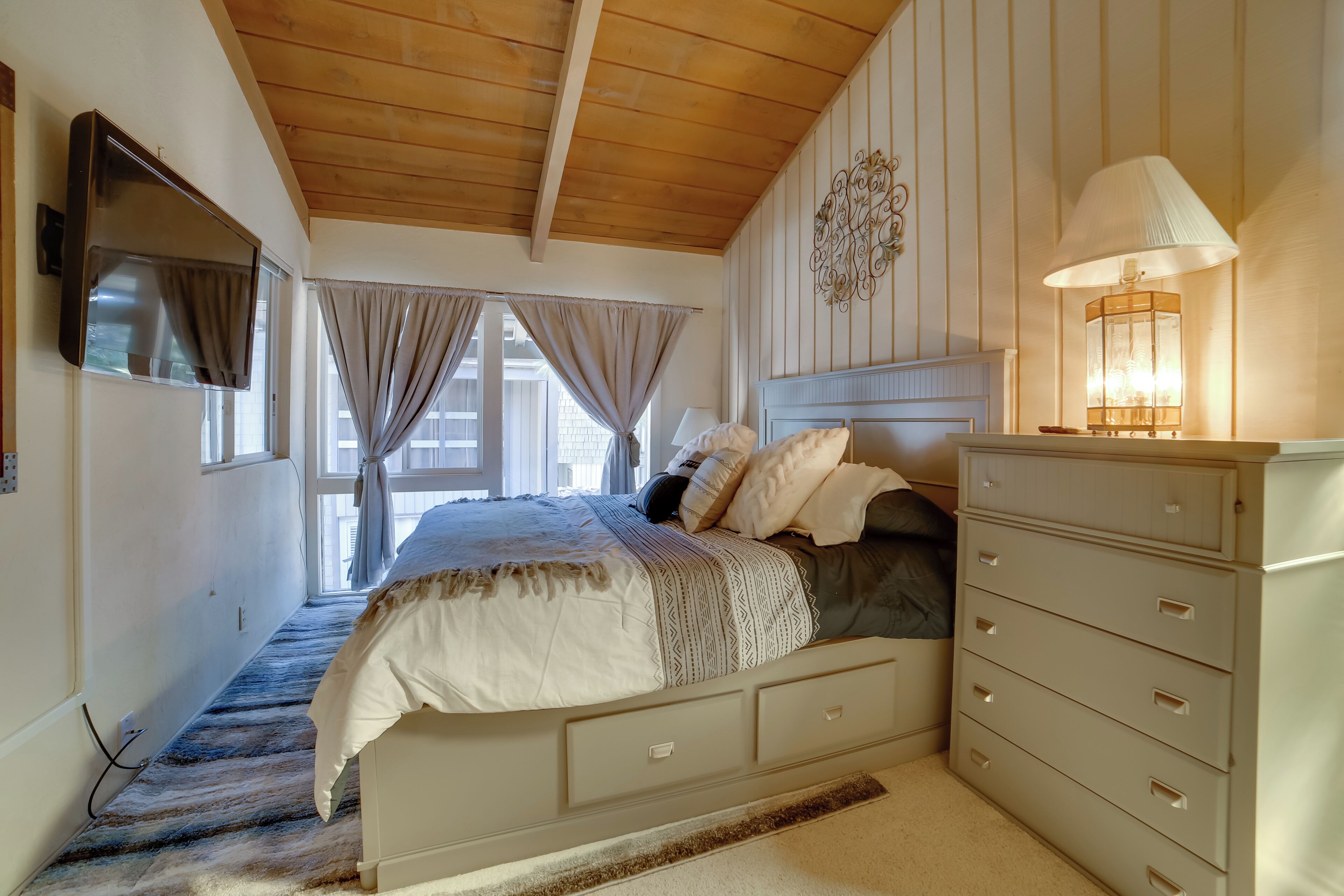 A bedroom with a bed, a dresser, a TV mounted on the wall, and a window with curtains.