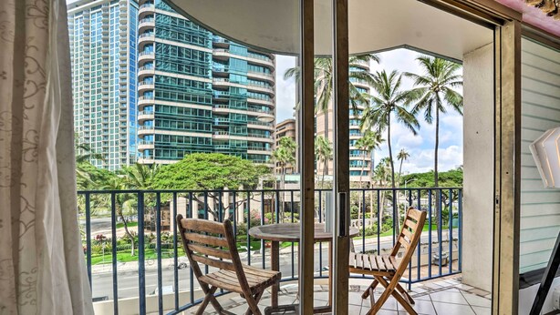 Un balcon avec deux chaises en bois et une petite table, donnant sur une rue bordée de palmiers et sur un grand immeuble.