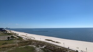 A coastal beach scene with a clear sky, a sandy beach, a pier extending into the water, and a road leading to the beach.
