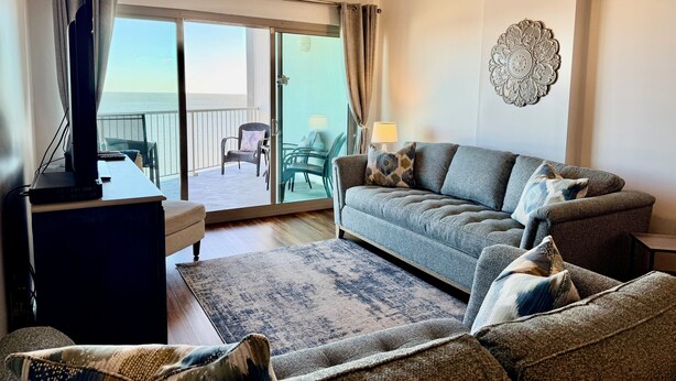 A living room with a sofa, a television, a ceiling fan, and a view of the ocean through a sliding glass door.