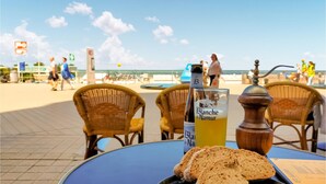 A table with a bottle of Blanche de Narmer, a glass of yellow drink, and a plate of bread at a beachside cafe with wicker chairs and a view of the ocean. - Lovely apartment in Oostende with WiFi (Oostende)