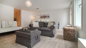 A modern living room with a grey sofa, ottoman, and a dining area with a table and chairs. There is a large window on the right, a painting on the wall, and a decorative light fixture. - Lovely apartment in Oostende with WiFi (Oostende)