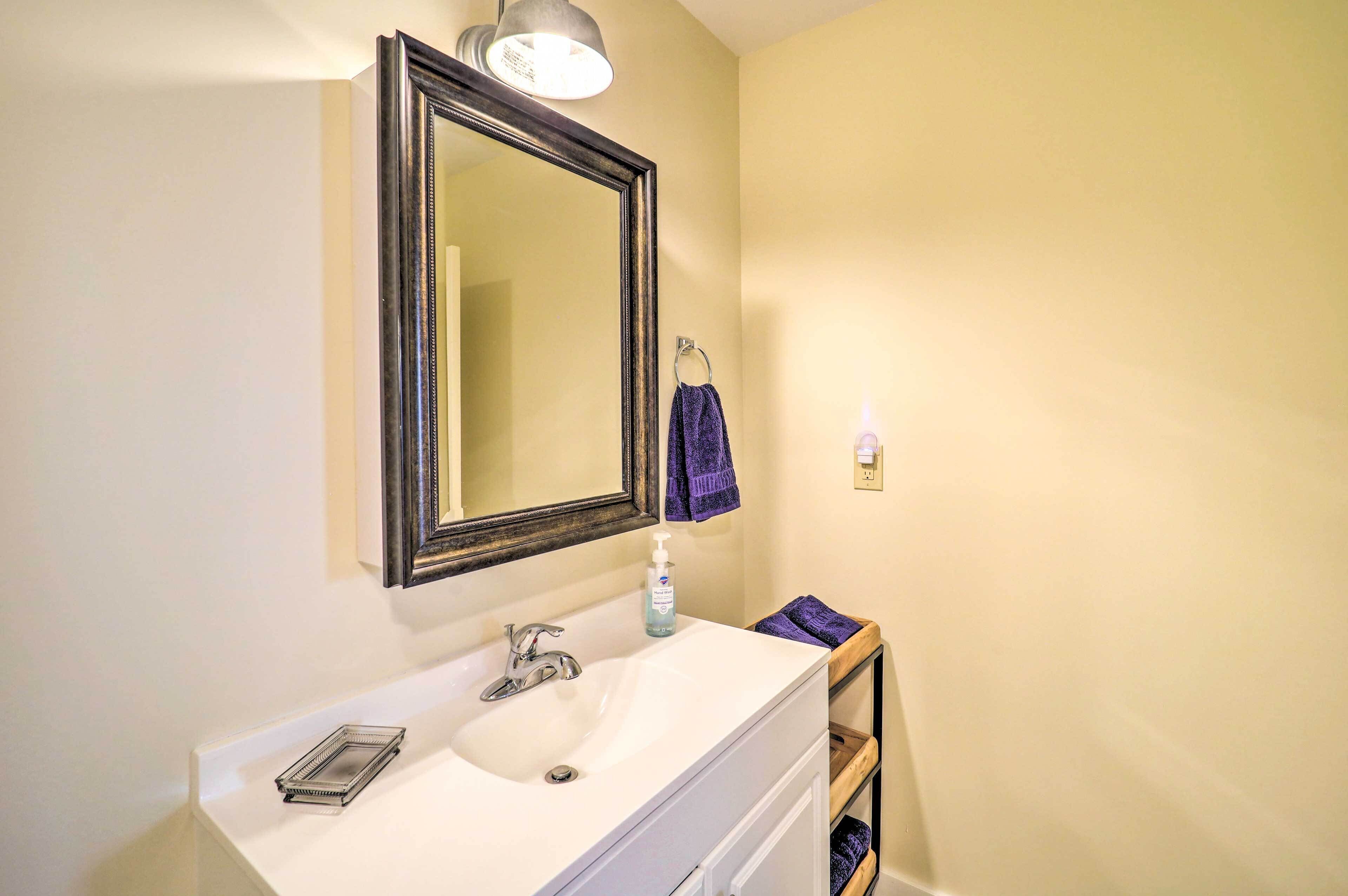 A bathroom with a white sink, a large mirror, a towel rack with a purple towel, and a soap dispenser.