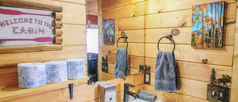 A wooden cabin bathroom with a sink, toilet paper, towels, and a welcome sign.