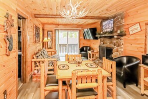 Een houten blokhut van binnen met een eettafel, stoelen, een stenen openhaard en een aan de muur bevestigde televisie.
