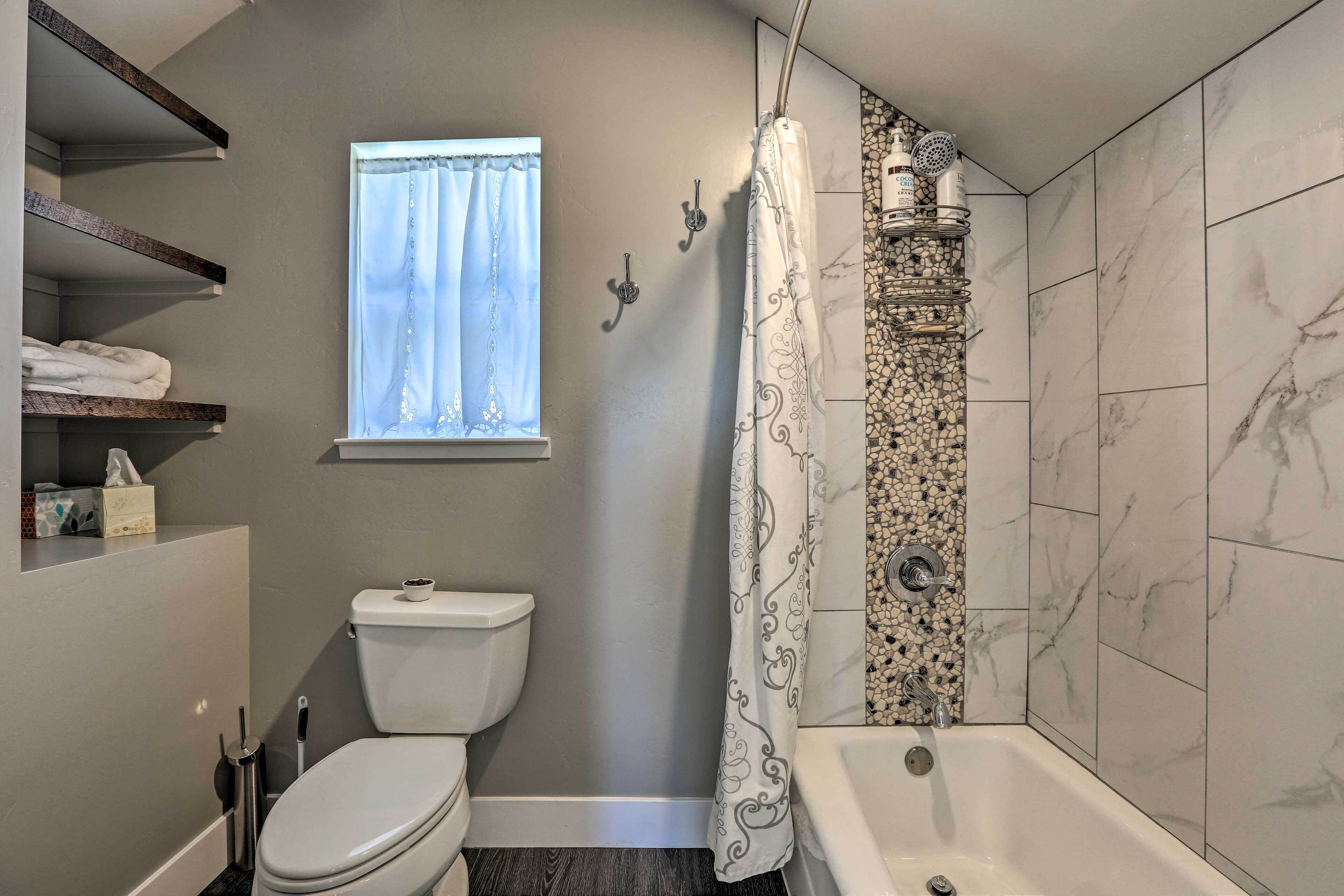 A bathroom with a toilet, a shower with a patterned curtain, a bathtub, and a window with a curtain. Shelves with towels and toiletries are visible.