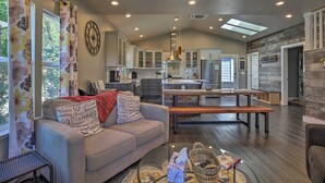 A modern living room with a sofa, a glass coffee table, and a bench. There is a kitchen in the background with white cabinets and a large clock. - Sierra Sunset Inn - 3 Miles to Placerville! (Diamond Springs)