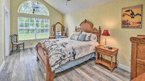 A bedroom with a large bed, a ceiling fan, a window with a view of greenery, a wooden nightstand, and a vintage poster on the wall. - Family Home: Walk to Downtown & Stetson Univ! (DeLand)