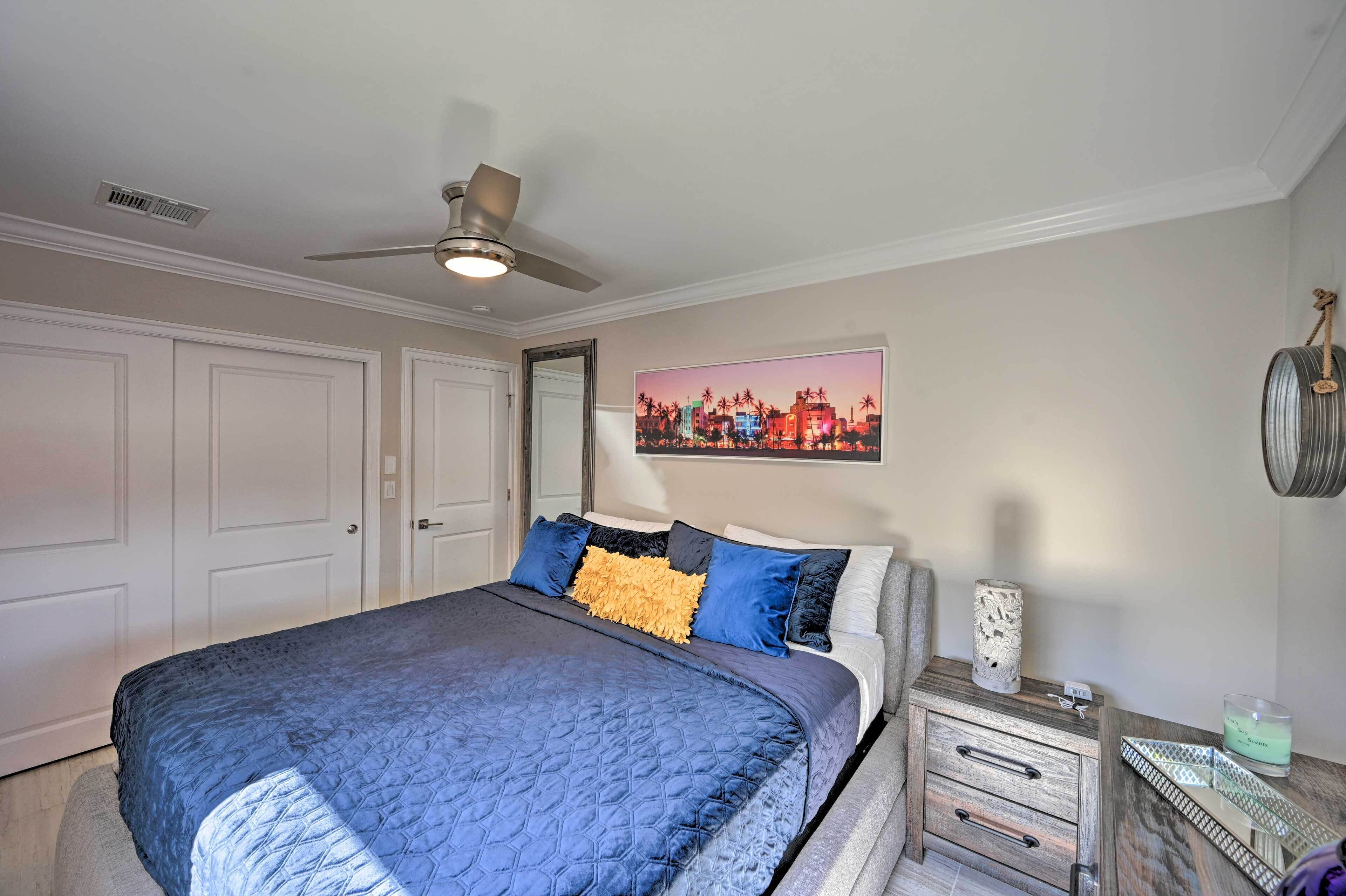 Une chambre à coucher comprenant un lit, une table de chevet, un ventilateur de plafond, un tableau accroché au mur et une porte.