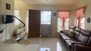 Una sala de estar con un sofá de cuero marrón, una mesa de comedor blanca con sillas, un televisor montado en la pared y una escalera que sube a otro nivel.