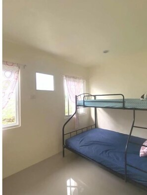 Quarto pequeno e único, com cama de solteiro, duas janelas com cortinas e uma parede branca simples.