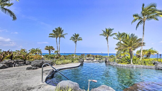 A clear blue pool with a stone deck, surrounded by palm trees and rocks, overlooking the ocean.