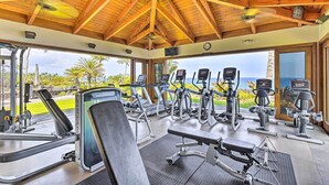 A gym with treadmills, elliptical machines, and weight benches, featuring a wooden ceiling and a view of the ocean.