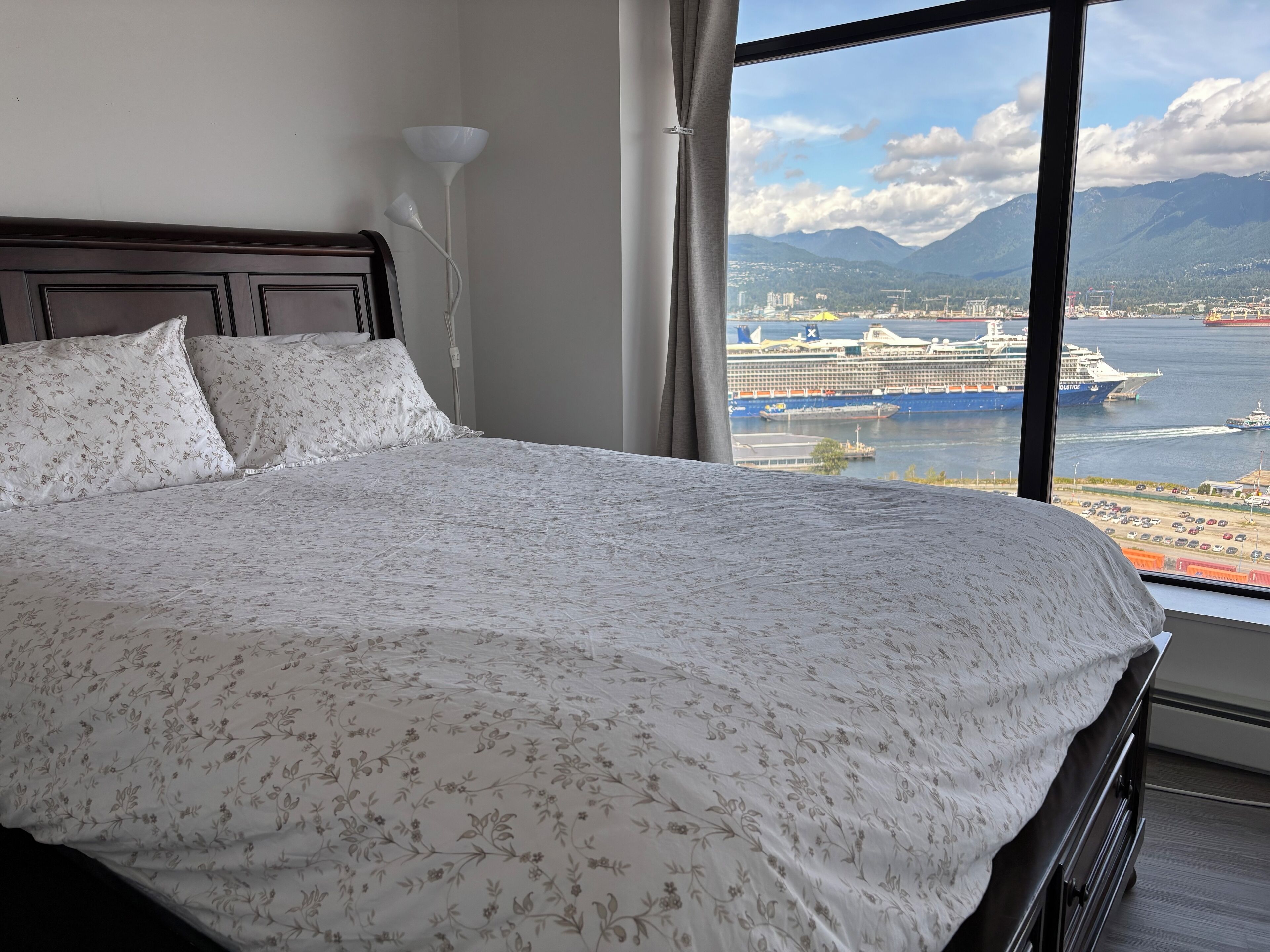 Una cama con ropa de cama floral, una lámpara de noche y vistas a un crucero y montañas a través de una ventana.