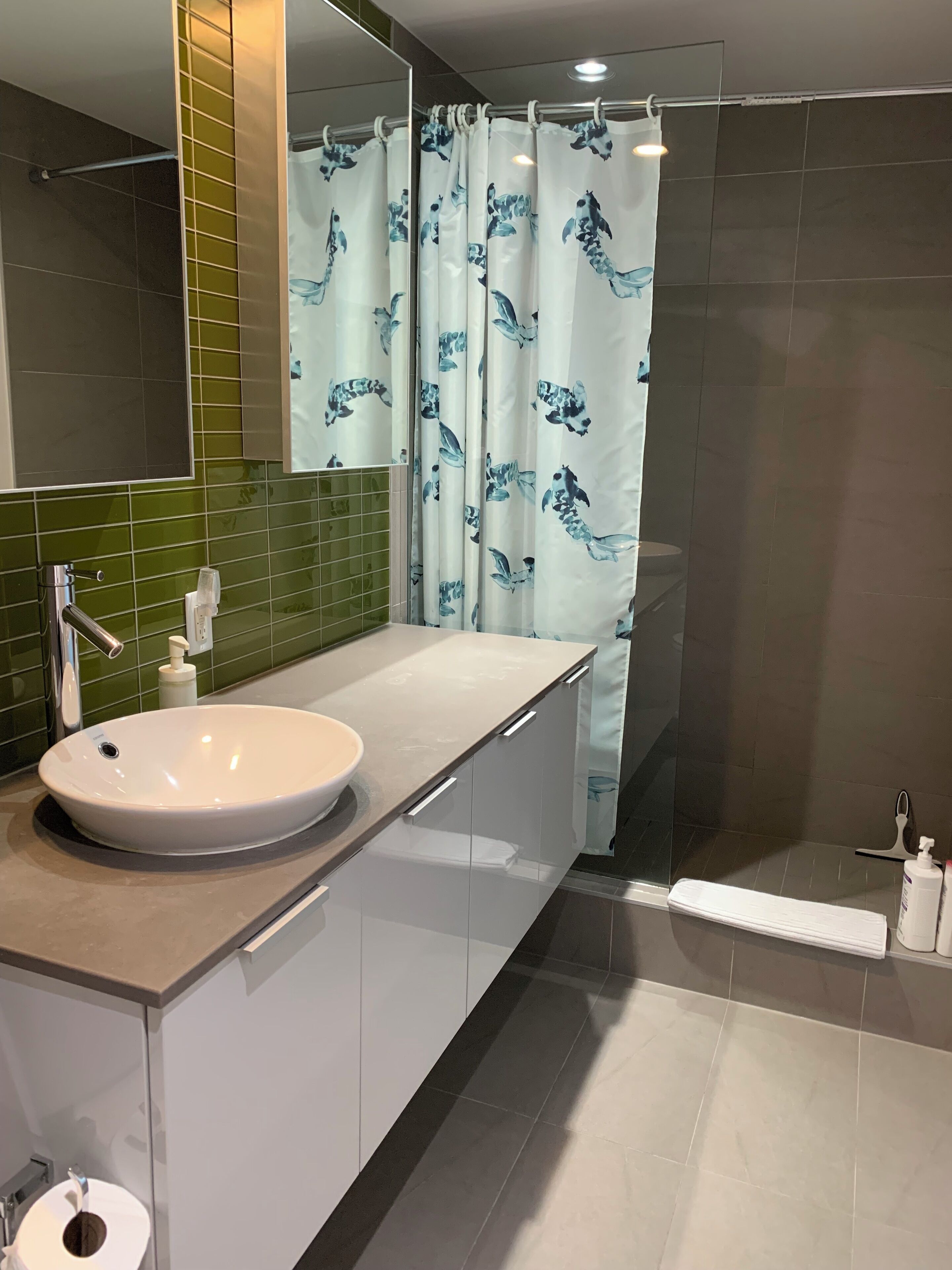 A modern bathroom with a white sink, green tiled walls, a shower curtain with a blue fish pattern, and a mirror above the sink.