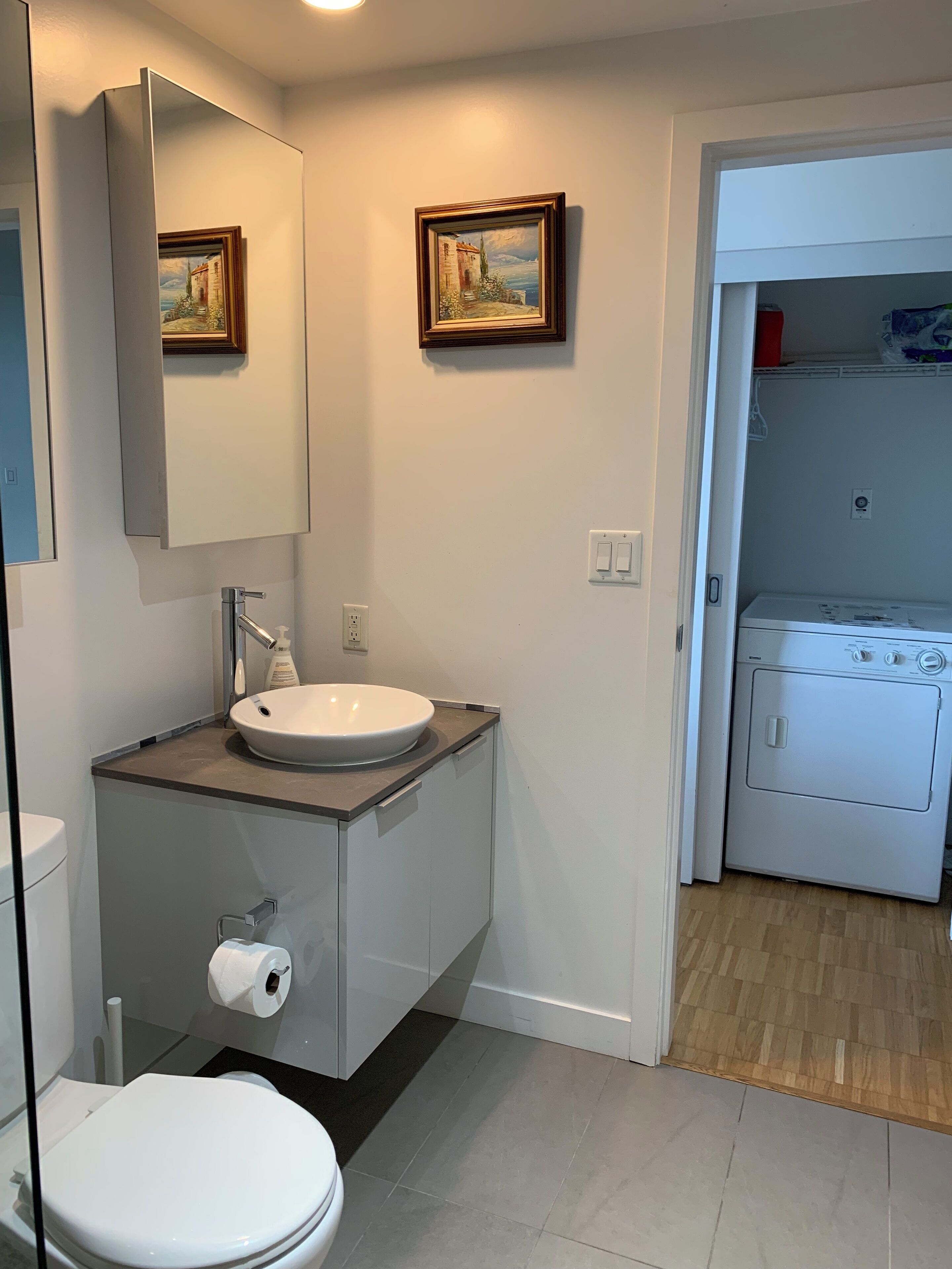 Ein modernes Badezimmer mit einem weißen Waschbecken, einem Spiegel und einer Toilette. An der Wand hängen zwei Bilder, und im angrenzenden Raum ist eine Waschmaschine zu sehen.