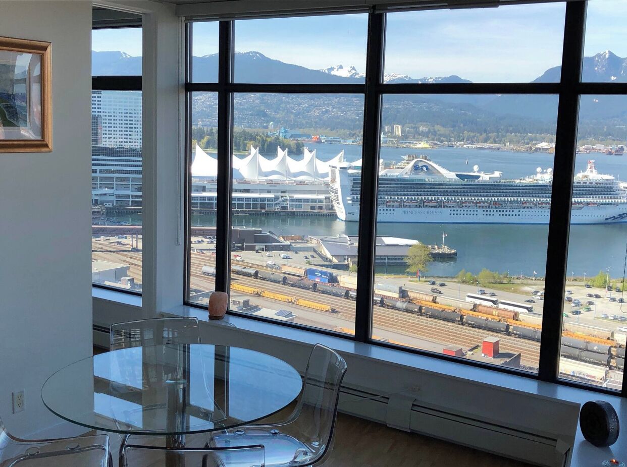 A room with a large window offering a view of a cruise ship, a train, and a cityscape with mountains in the background.