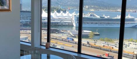 A room with a large window offering a view of a cruise ship, a train, and a cityscape with mountains in the background.
