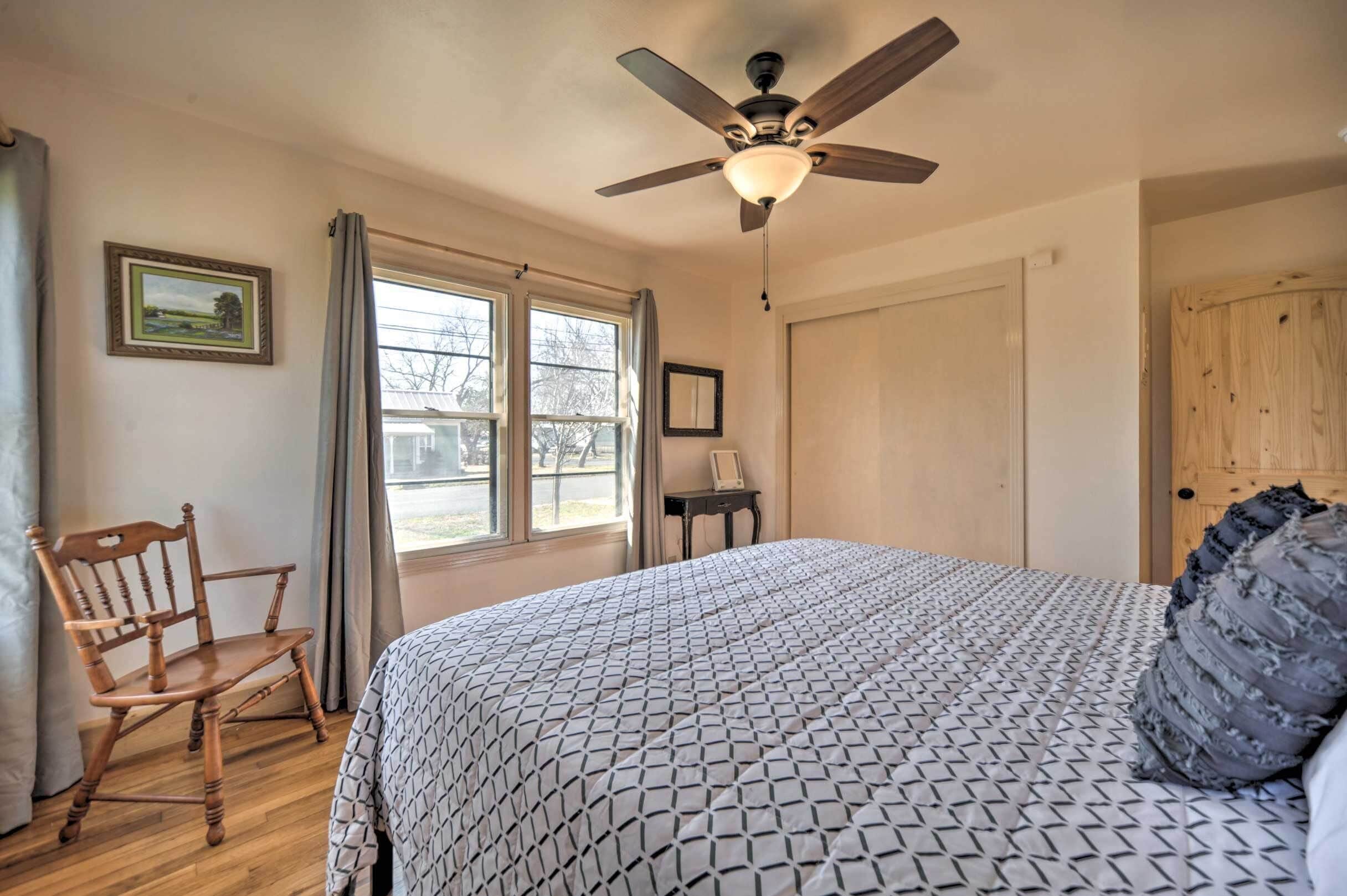 Un dormitorio con cama, una silla de madera, un ventilador de techo y una ventana con cortinas.