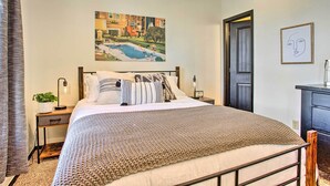 A neatly made bed with a patterned blanket, a nightstand with a lamp, a framed picture on the wall, and a door to another room. - Norman, Oklahoma, Vacation Rental 1 Mi to OU! (Norman)