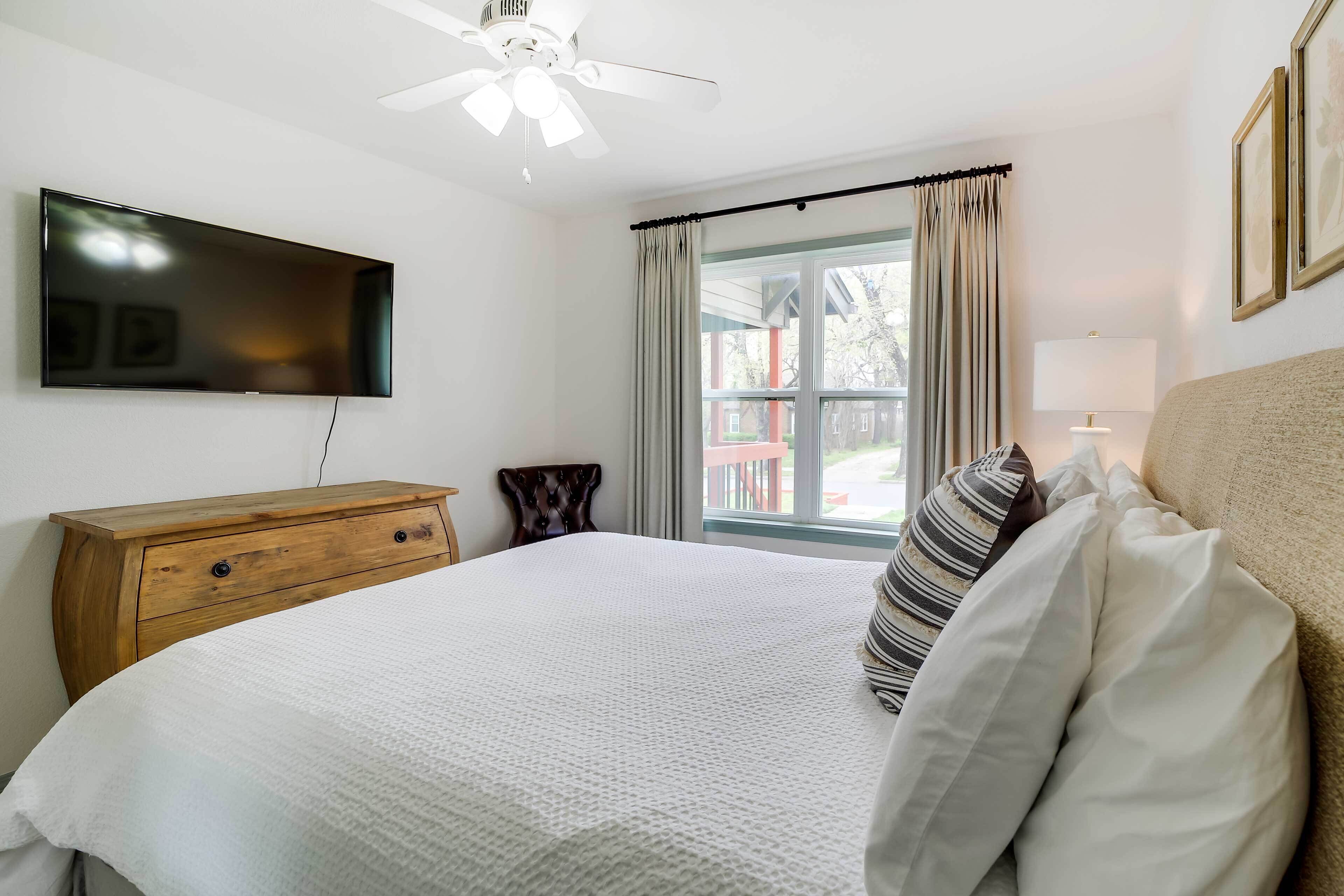 A bedroom with a bed, a wooden dresser, a flat-screen TV, a ceiling fan, a window with curtains, and a lamp.