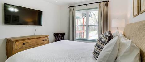 A bedroom with a bed, a wooden dresser, a flat-screen TV, a ceiling fan, a window with curtains, and a lamp.