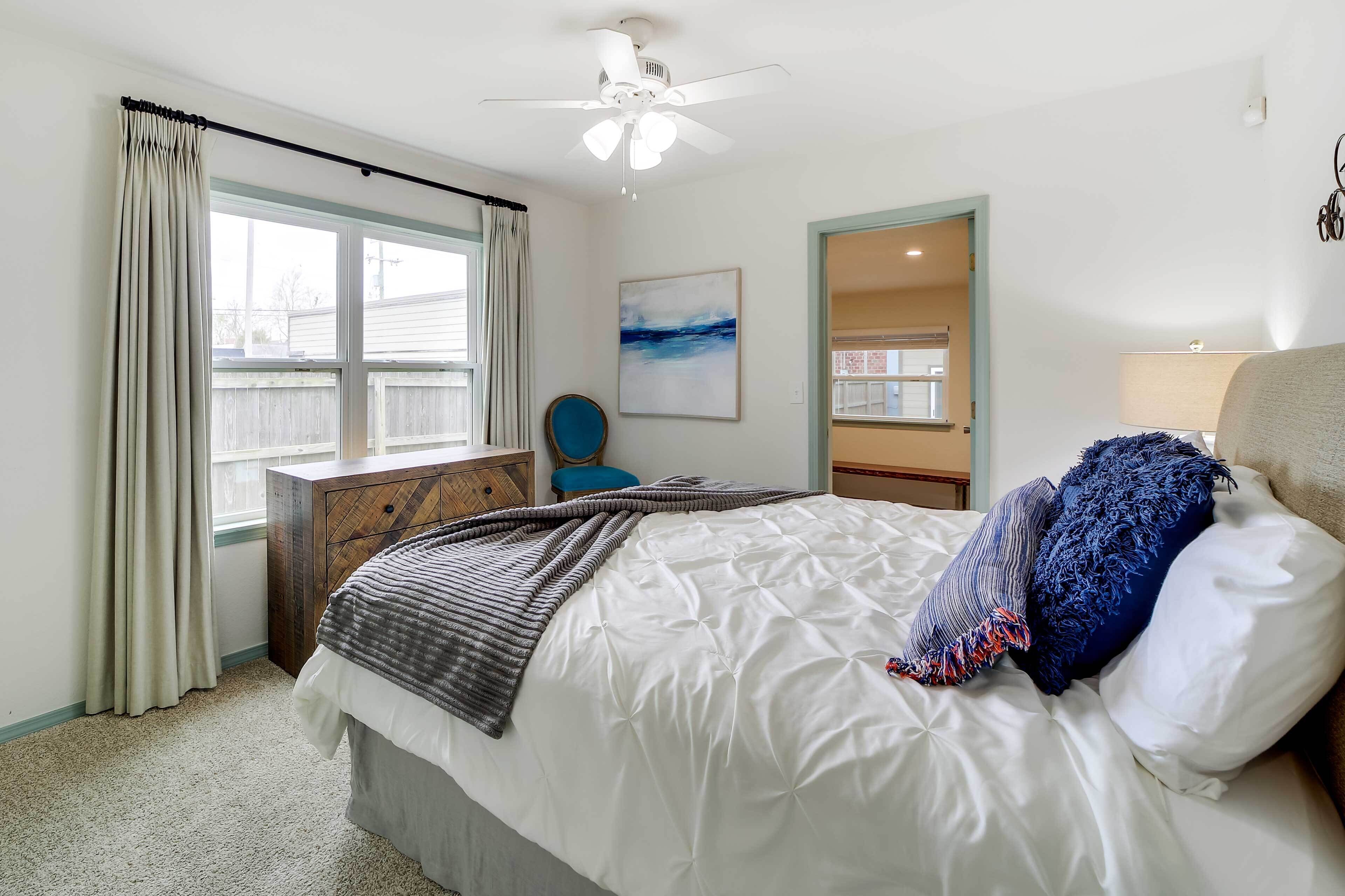 A bedroom with a bed, a ceiling fan, a window with curtains, a chair, and a painting on the wall.