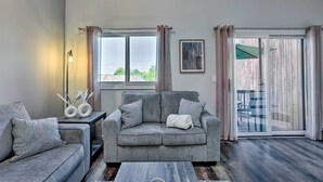 Un salón con un sofá gris, una mesa de centro y una mesita de noche con objetos decorativos. Se ve una ventana con cortinas y vistas al exterior.