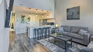 Una sala de estar moderna con un sofá gris, una mesa de centro y una zona de cocina con taburetes de bar.