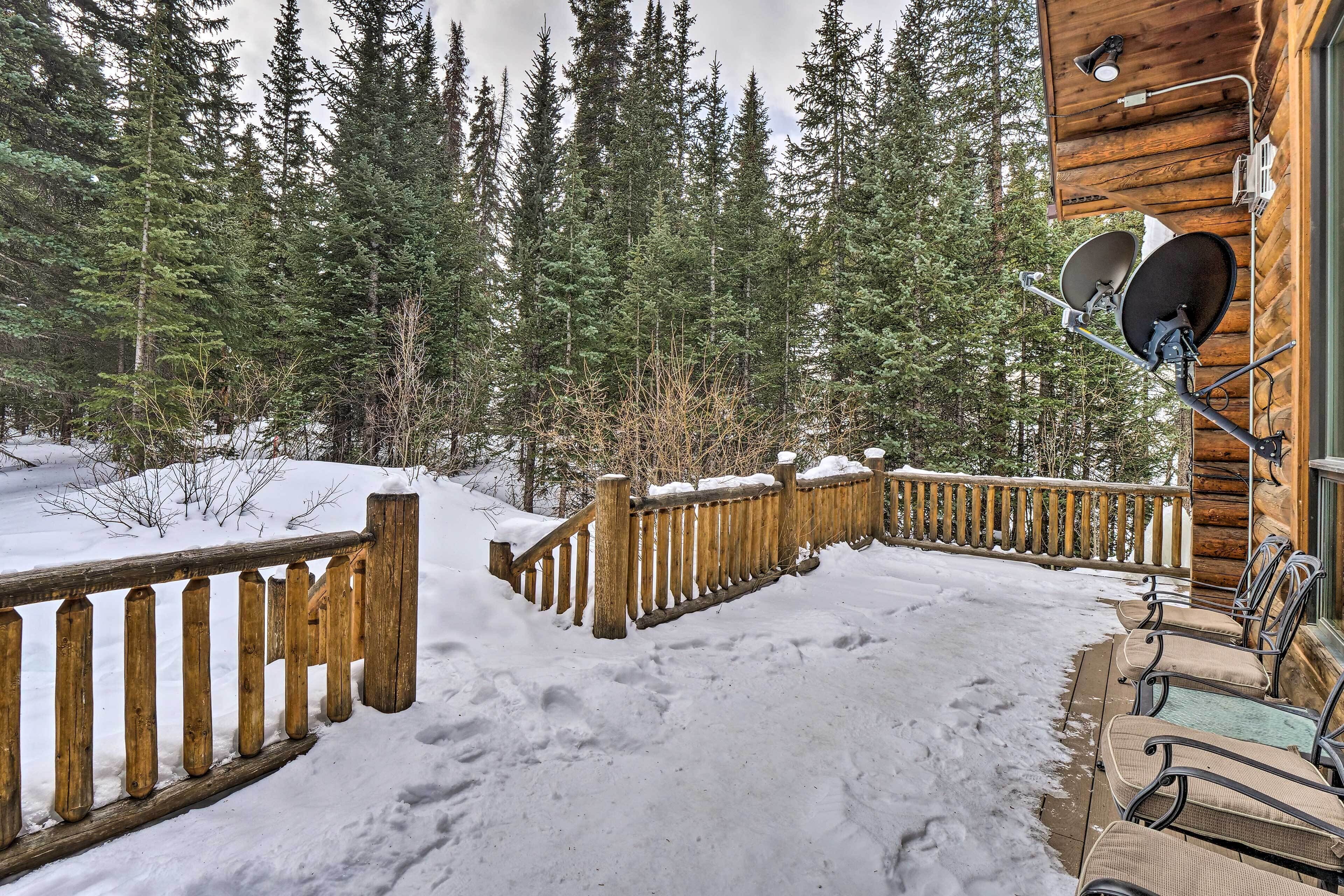 A wooden cabin with a deck, satellite dishes, and a snowy forest backdrop.