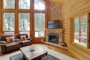 L’intérieur d’une cabane en rondins, avec une cheminée en pierre, un fauteuil inclinable en cuir et de grandes fenêtres donnant sur une forêt.