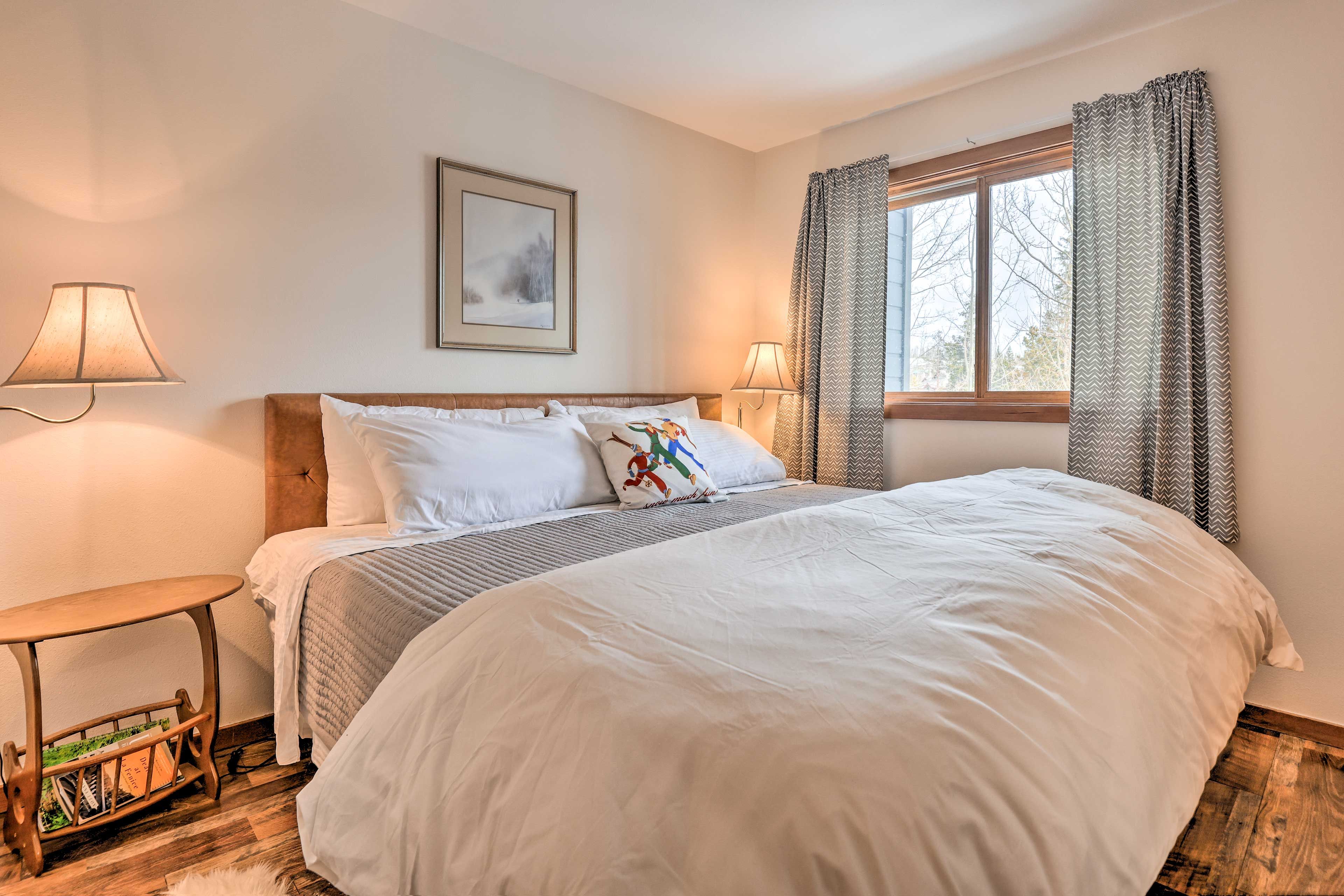 A neatly made bed with white bedding, two bedside lamps, a framed picture on the wall, and a window with curtains.
