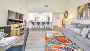 Una sala de estar moderna con un sofá gris, una mesa de centro de vidrio y una alfombra colorida. Se observa una cocina al fondo con un taburete de bar y un televisor montado en la pared.