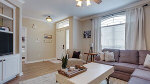 A spacious living room with a large sectional sofa, a coffee table, and a TV. There is a door leading to another room and a window with blinds.