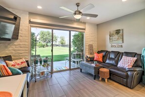 Un salon avec un canapé en cuir, une table basse et un téléviseur fixé au mur. On y trouve également un ventilateur de plafond, une porte-fenêtre coulissante donnant sur un patio, ainsi qu’une petite table avec des chaises à l’extérieur.