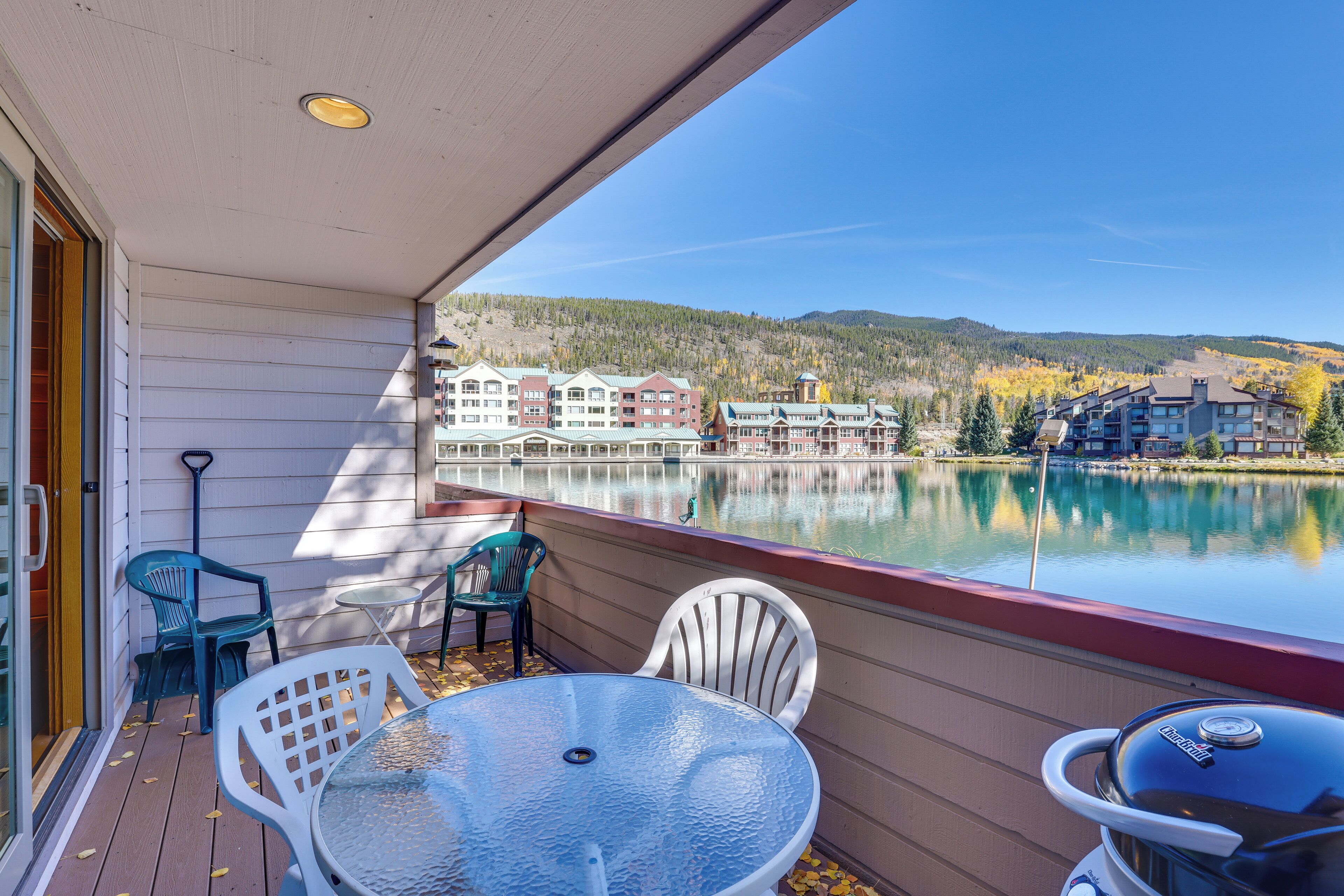 A balcony with a glass table and chairs, overlooking a lake and buildings.