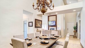 Un coin repas est aménagé avec une longue table en bois, dressée pour six personnes, et entourée de chaises rembourrées. Un lustre est suspendu au plafond et un téléviseur est fixé au mur.