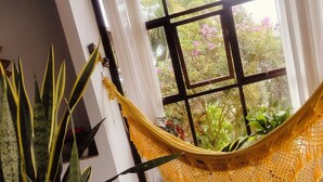 Una habitación con una hamaca, una planta en maceta, una ventana con vistas a la vegetación y una mesa blanca.